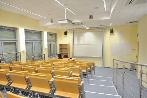 Sala 323 - amfiteatralne rzędy krzeseł z pulpitami, schody z poręczą i ekran projekcyjny.