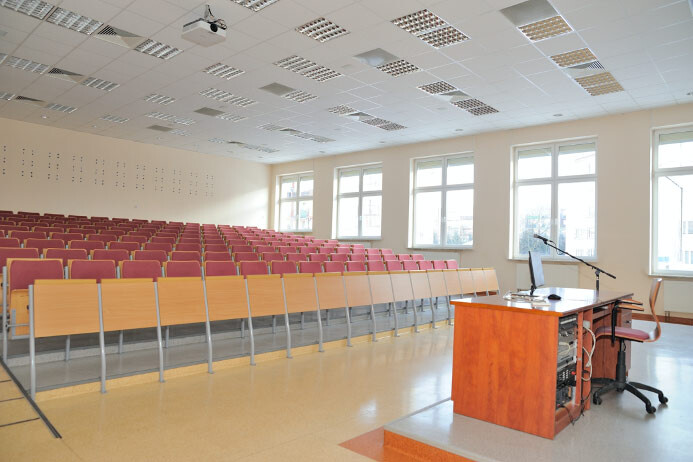 Sala B107 - stanowisko prowadzącego z komputerem i mikrofonem na tle rzędów siedzeń.