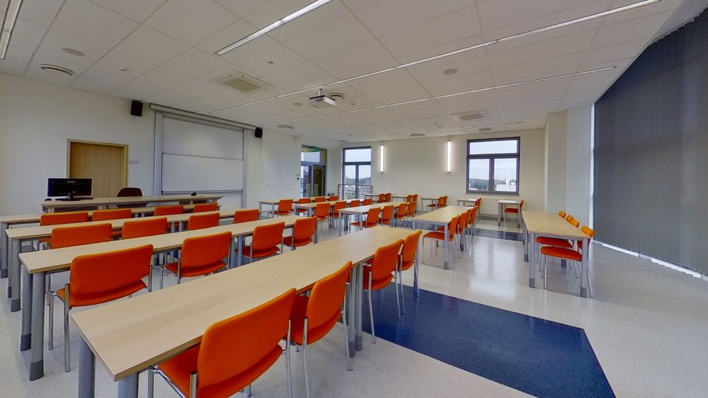 Sala A423 - widok od strony okien na rzędy stołów, stanowisko prowadzącego i ekran.