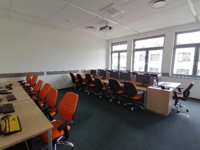 Pracownia A204 - stanowiska komputerowe wzdłuż ścian, pomarańczowe krzesła i projektor sufitowy.